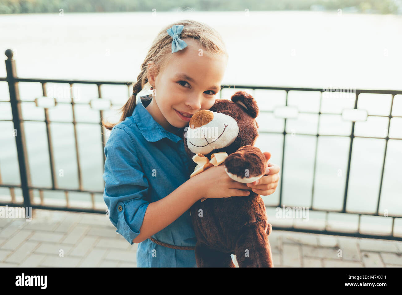 Close-up portrait of girl embracing a cute teddy bear looking to camera on a lake with trees background wearing denim dress. Kids friendship concept.  Stock Photo