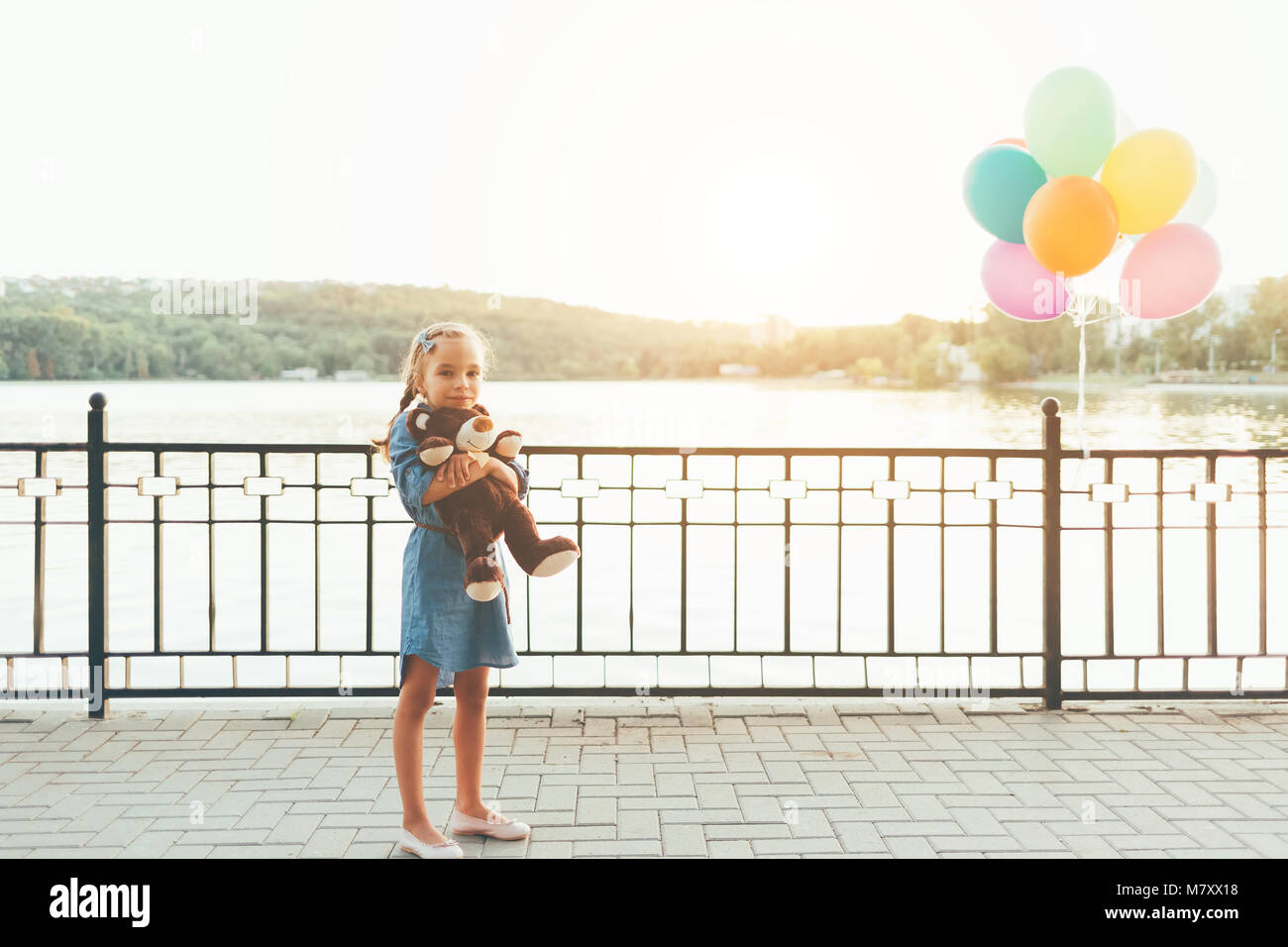 Full length portrait of girl hugging a cute teddy bear looking to camera on a lake with trees and balloons background wearing denim dress. Kids friend Stock Photo