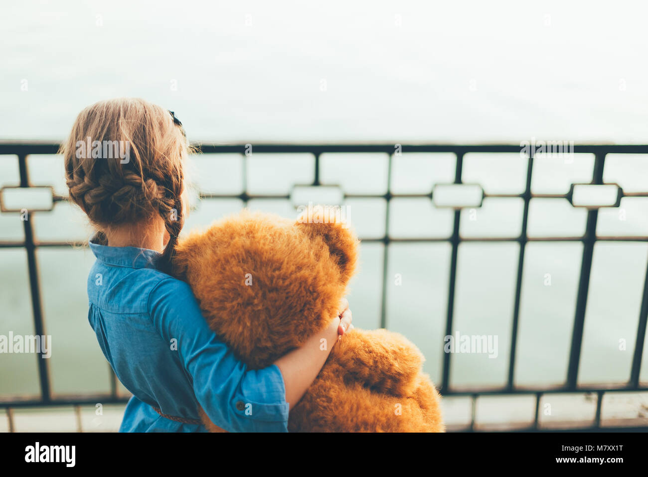 Back view of girl embracing a cute teddy bear looking to lake wearing denim dress. Kids friendship concept. Friendly kid. Copy space available. Stock Photo