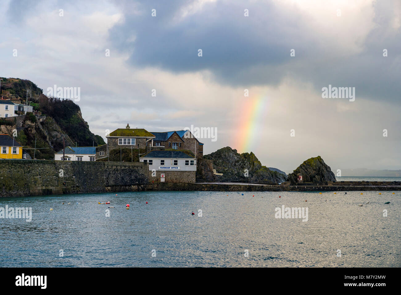A Large Rainbow Streaks into the Sky at Mevagissey Harbour in Cornwall. The Harbour offices Generic sign can be seen clearly. Stock Photo