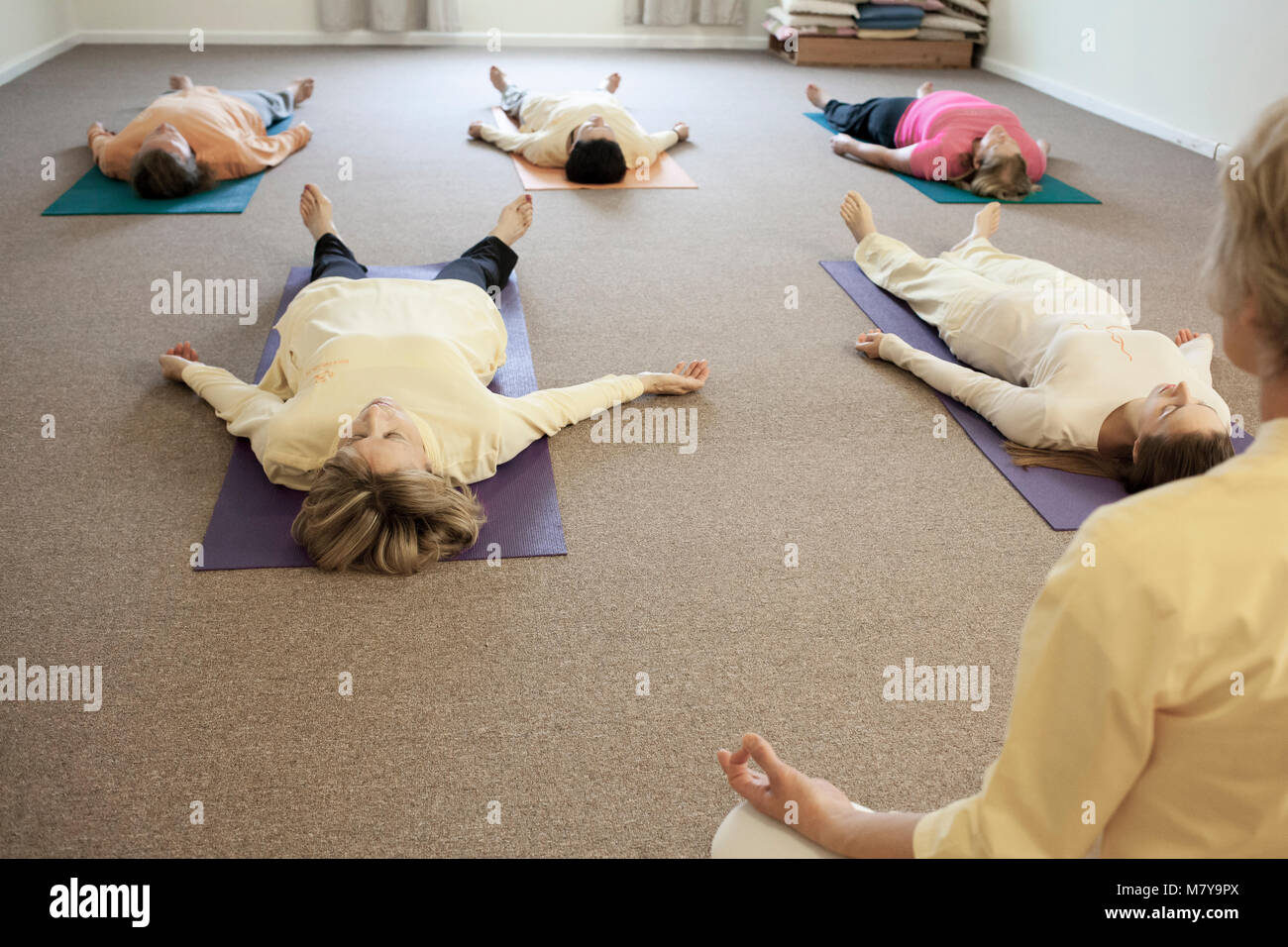 Young people practising relaxation also called yoga nidra. Guided by a certified female yoga instructor laying in shavasana on theri backs on yoga mat Stock Photo