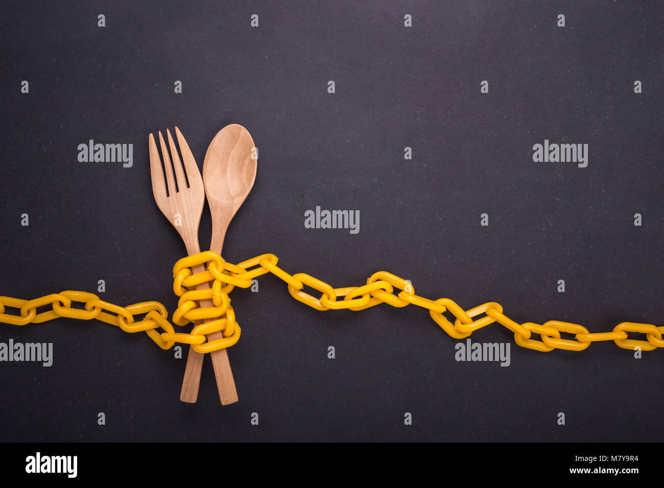 Yellow chain locked around the wooden spoon and fork on black stone board. Stop eating, eating control or diet concept. Top view Stock Photo