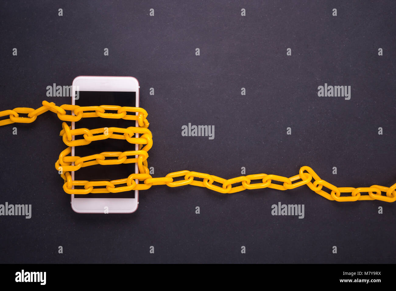 Yellow chain locked around the smartphone on black stone board. Stop, control or avoid using smartphone concept. Top view Stock Photo