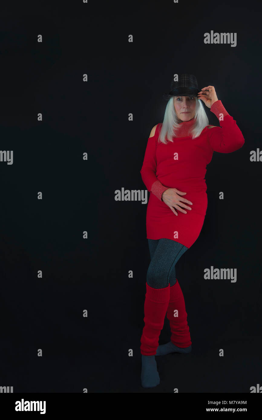 Portrait of a mature wearing gray and black hat on top of white hair, red sweater and leg warmers and gray leggings.  Posed in a minimalist style agai Stock Photo