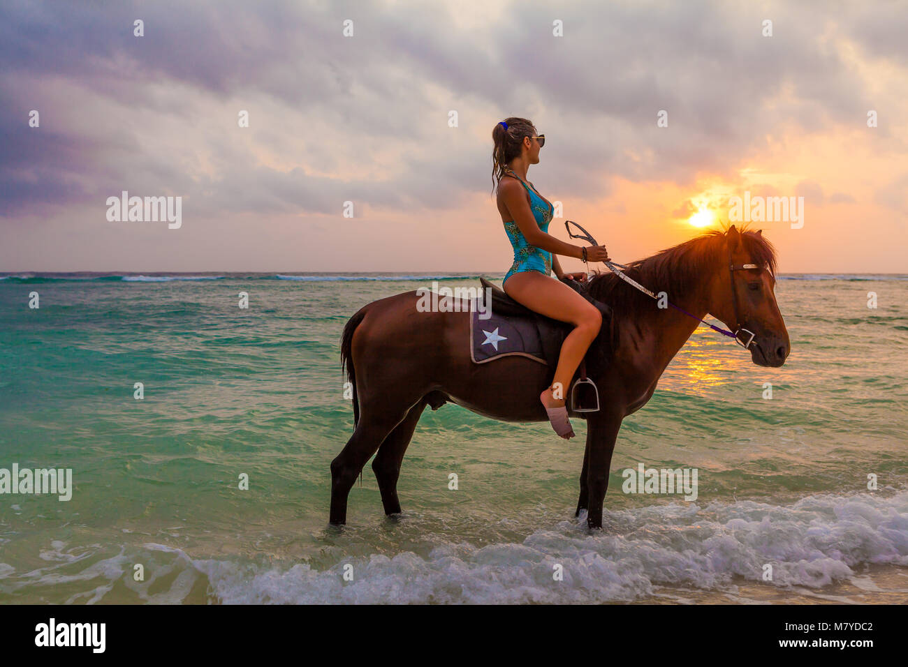 Beautiful girl riding a horse in the water, on the deserted beach of Gili Trawangan island, Indonesia, looking at the sunset over the sea Stock Photo
