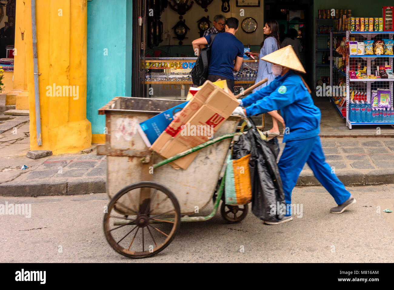 A street sweeper wearing a traditional bamboo conical hat, pushes a hand cart for collecting litter in Hoi An, Vietnam Stock Photo