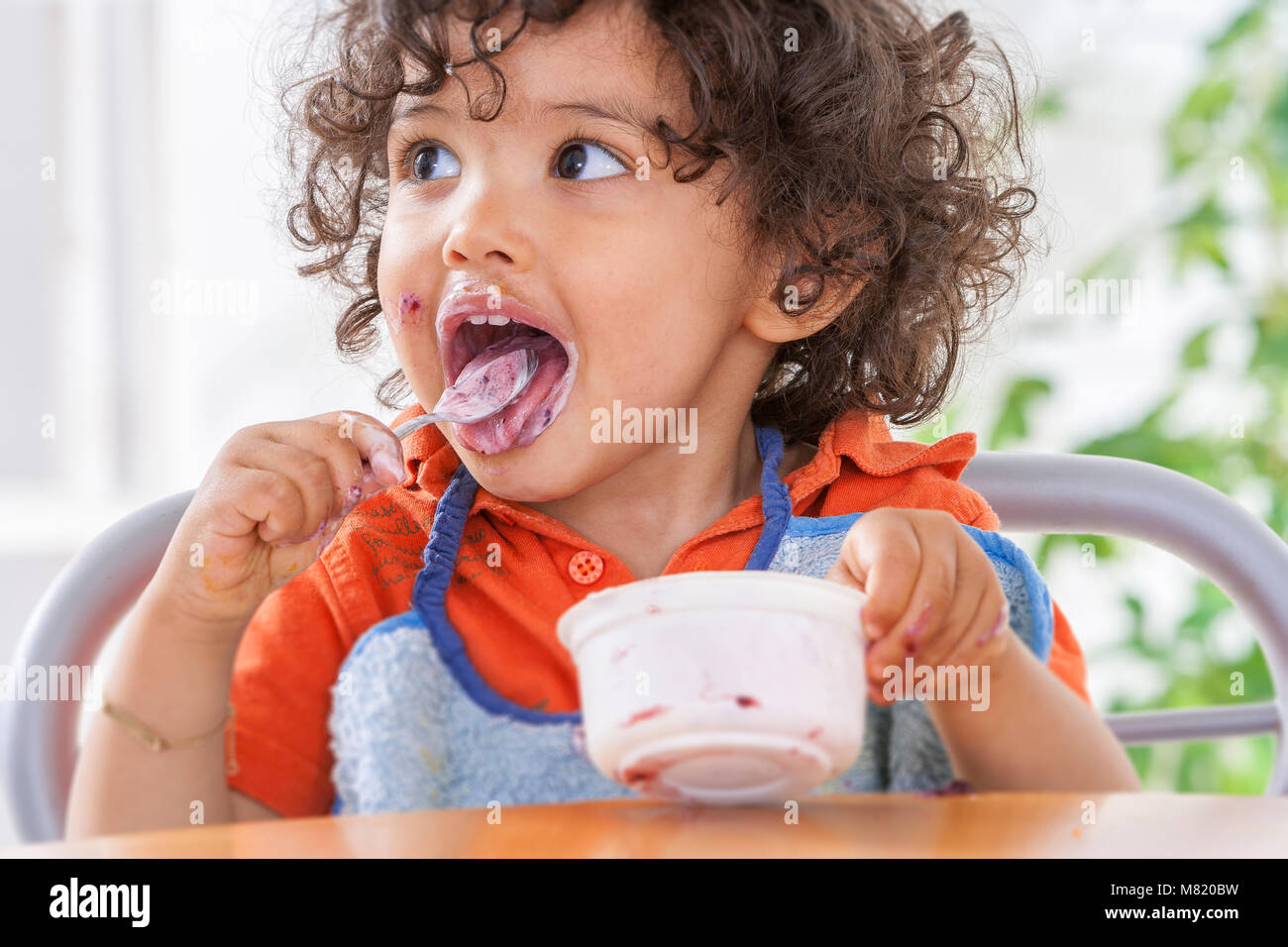 Toddler sitting in highchair and eating greek yogurt. Baby learning to eat and has yogurt on face and hair Stock Photo