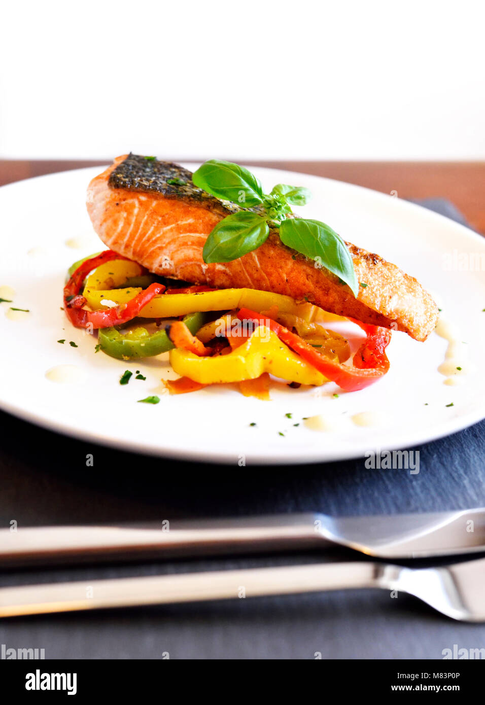Delicious salmon filet and red bell pepper vegetables on a white plate. Healthy meal, decorated with basil leaf. Dinner scene with white wine and fish. Stock Photo