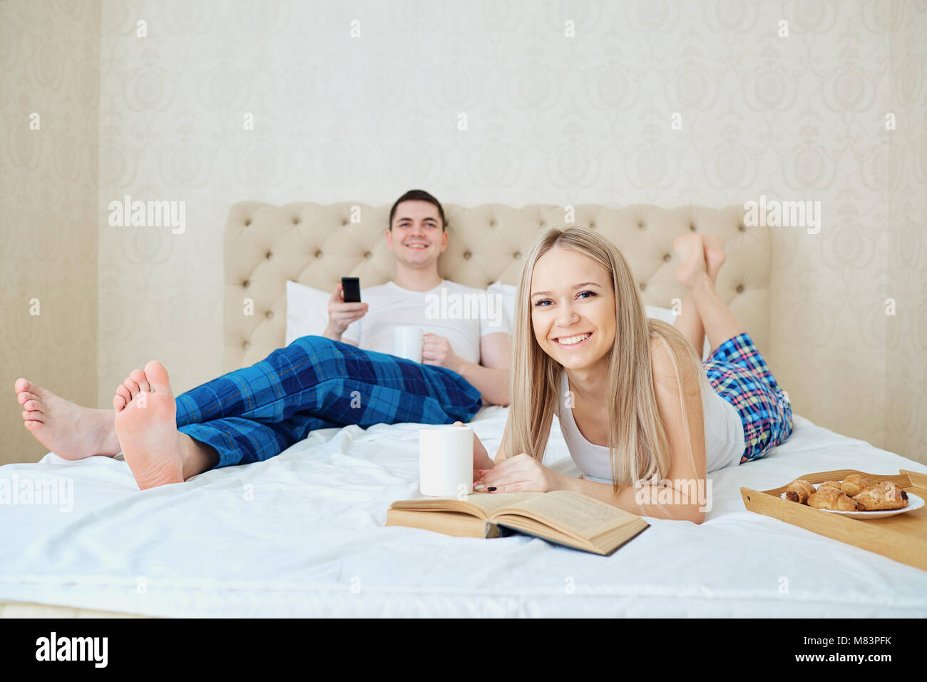 A couple in bed is having breakfast, watching TV, reading a book in the bedroom. Stock Photo