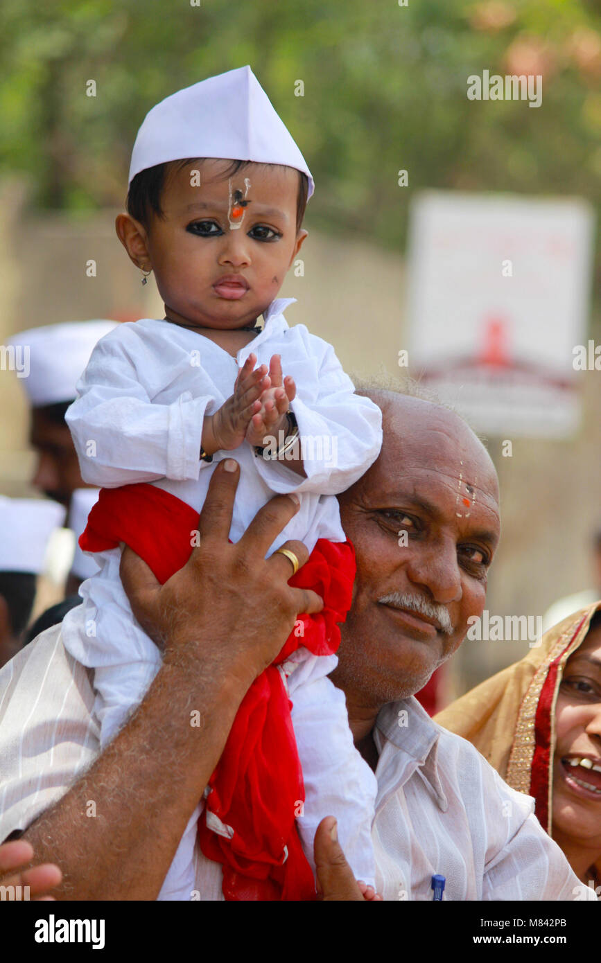 Young child with white cap and dress being carried on father's shoulders. Pandharpur festival Stock Photo