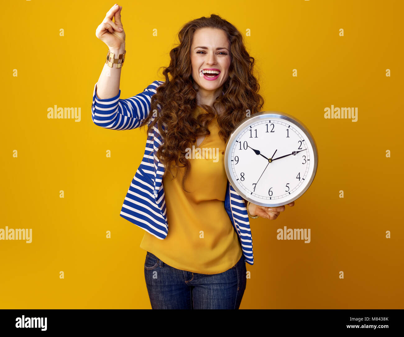 happy modern woman with long wavy brunette hair on yellow background with clock snapping fingers Stock Photo