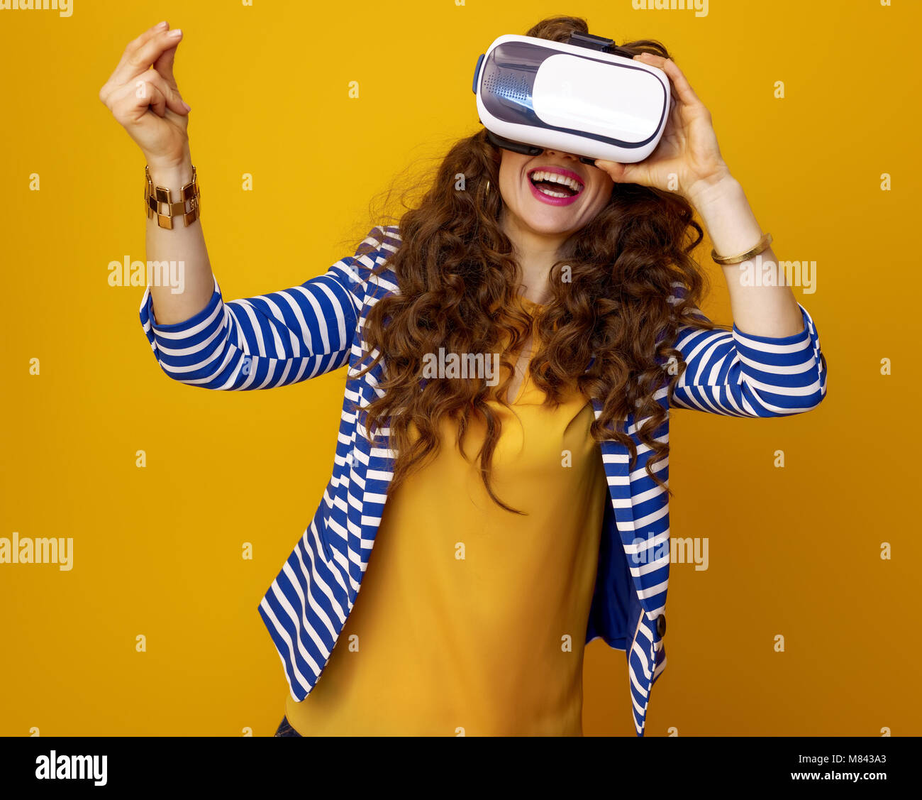 happy stylish woman with long wavy brunette hair against yellow background using virtual reality gear and snapping fingers Stock Photo