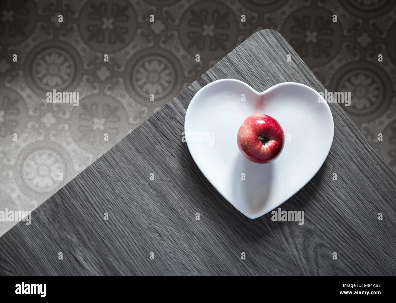 Looking down from above onto a table with a heart shaped plate and a colourful red apple in grey surroundings. Stock Photo