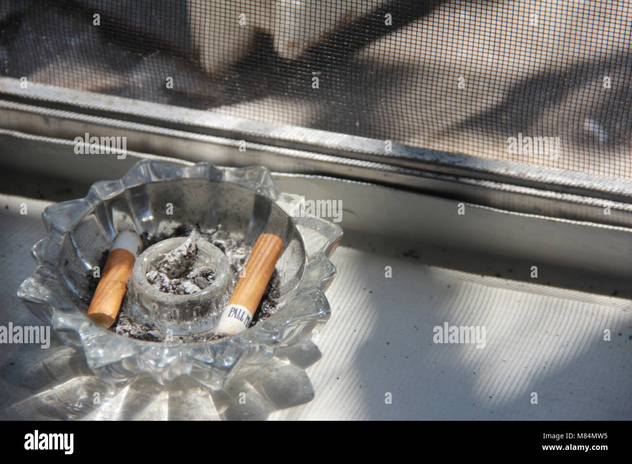 Cigarettes in a crystal ashtray on window sill. Stock Photo
