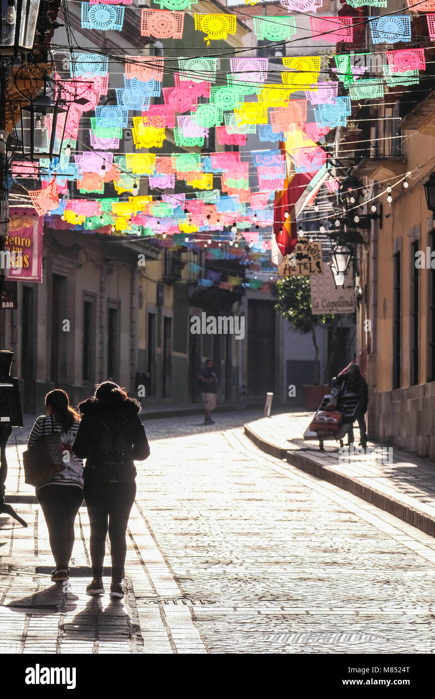 Mexicans walking on cobblestone street with sun lit colorful banner flags hanging above Stock Photo
