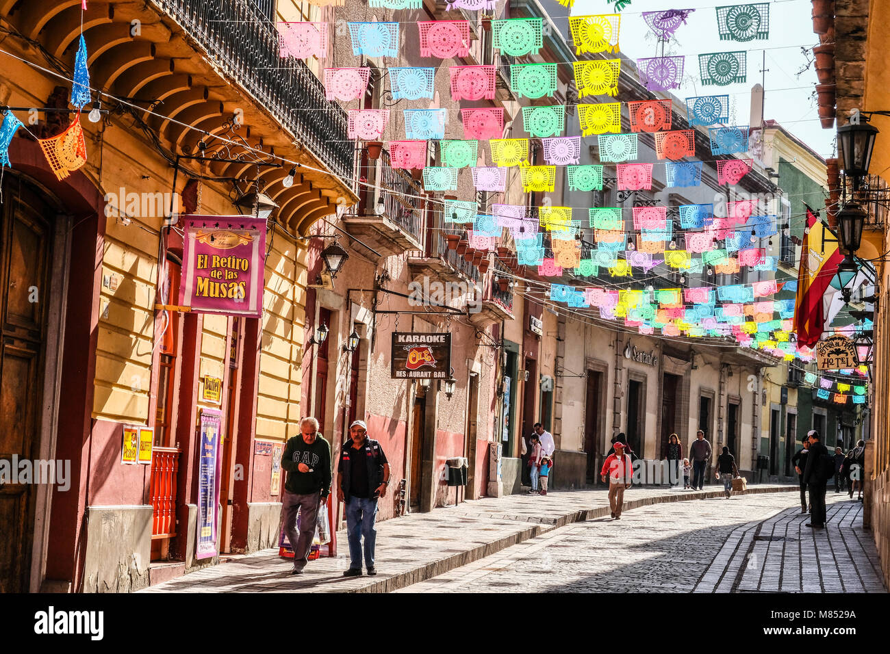 Mexicans walking on cobblestone street with sun lit colorful banner flags hanging above Stock Photo
