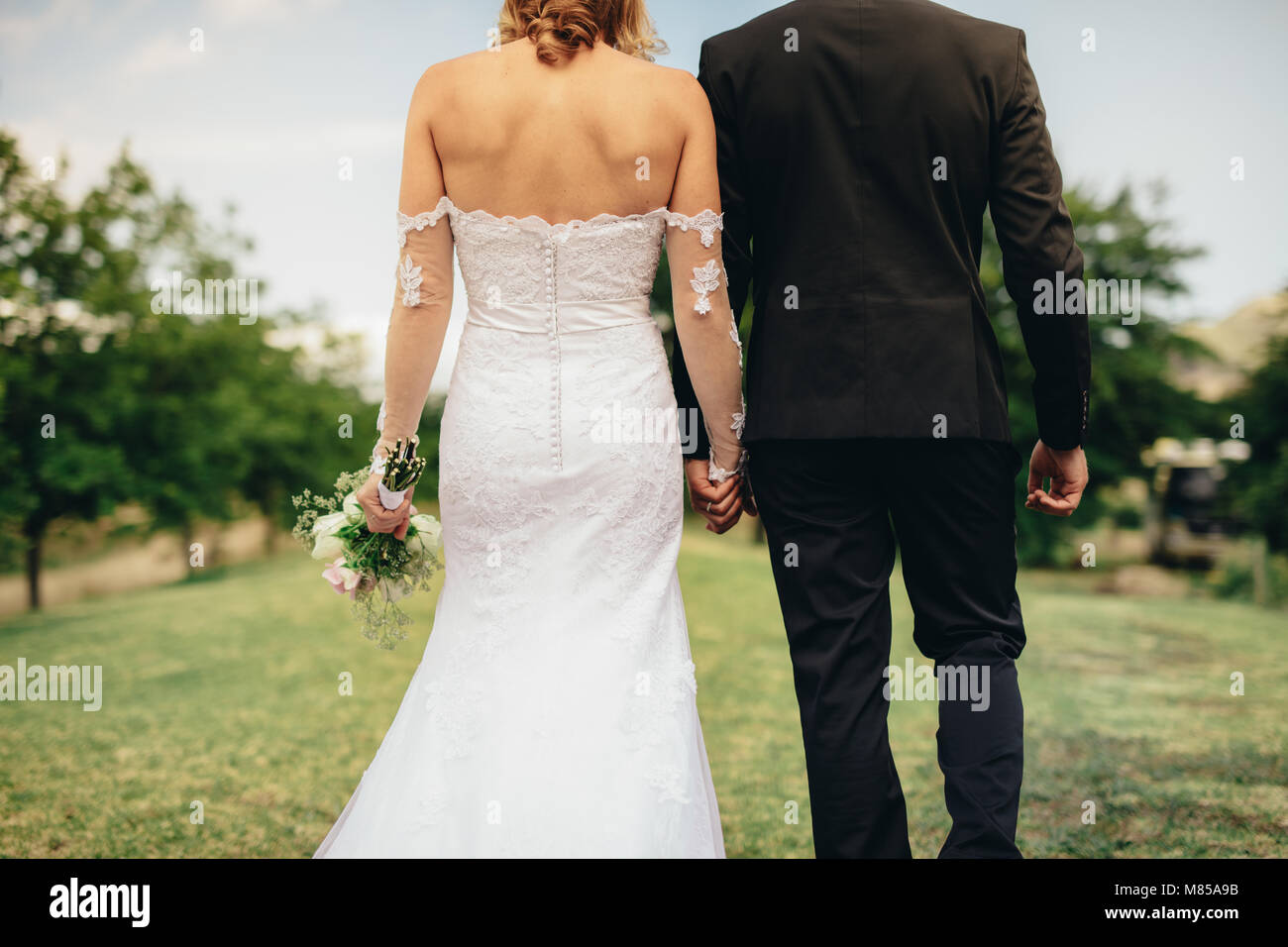 Rear view of bride and groom walking and holding hands on a meadow ...