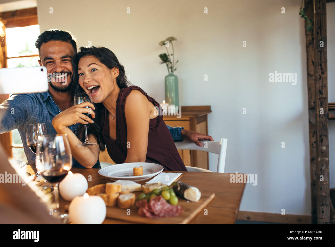 Young man and woman sitting at dining table laughing while taking selfie. Couple enjoying party at home. Stock Photo