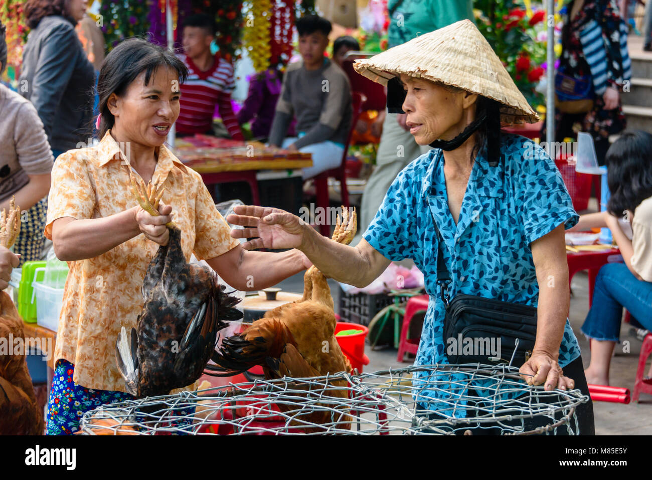 A customer laughs with a woman wearing a traditional Vietnamese conical bamboo hat as she holds a chcken by the legs in Hoi An, Vietnam Stock Photo