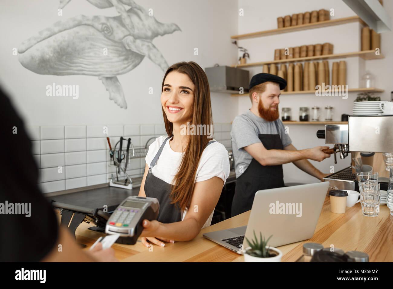 Coffee Business Concept - Beautiful female barista giving payment service for customer with credit card and smiling while working at the bar counter in modern coffee shop. Stock Photo