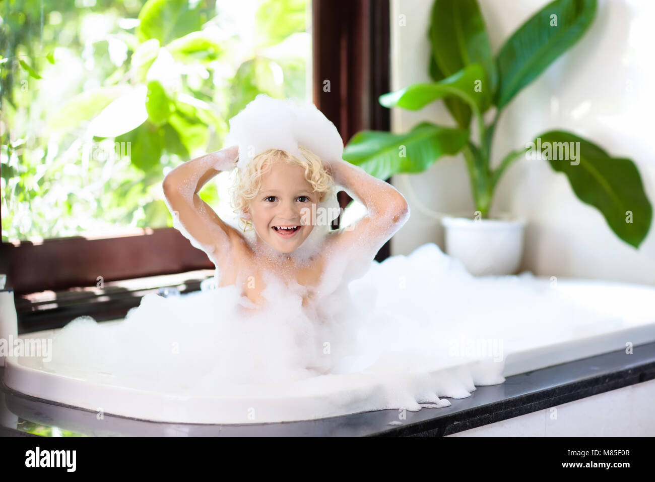 Little child taking bubble bath in beautiful bathroom with big garden