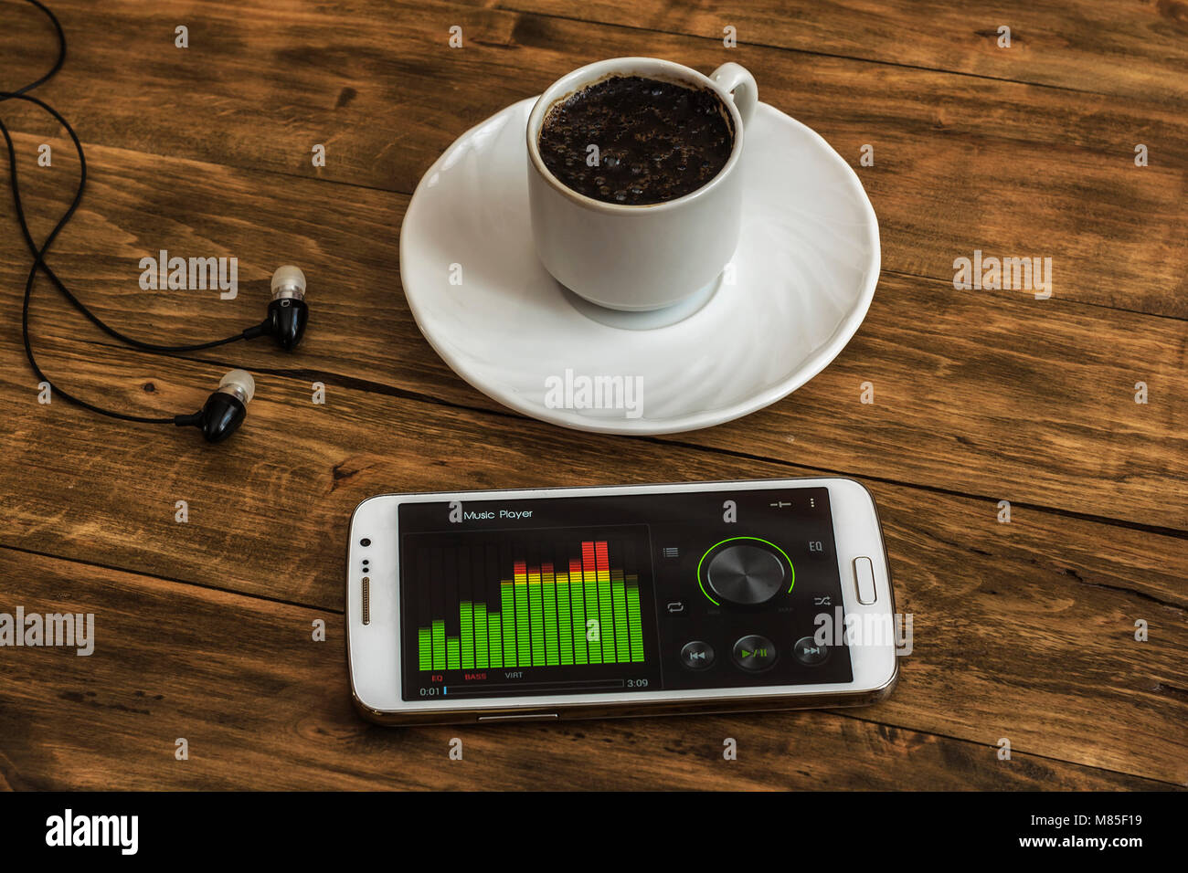 On the wooden surface there is a Cup of black coffee on a saucer, next are headphones and a smartphone with an equalizer Stock Photo