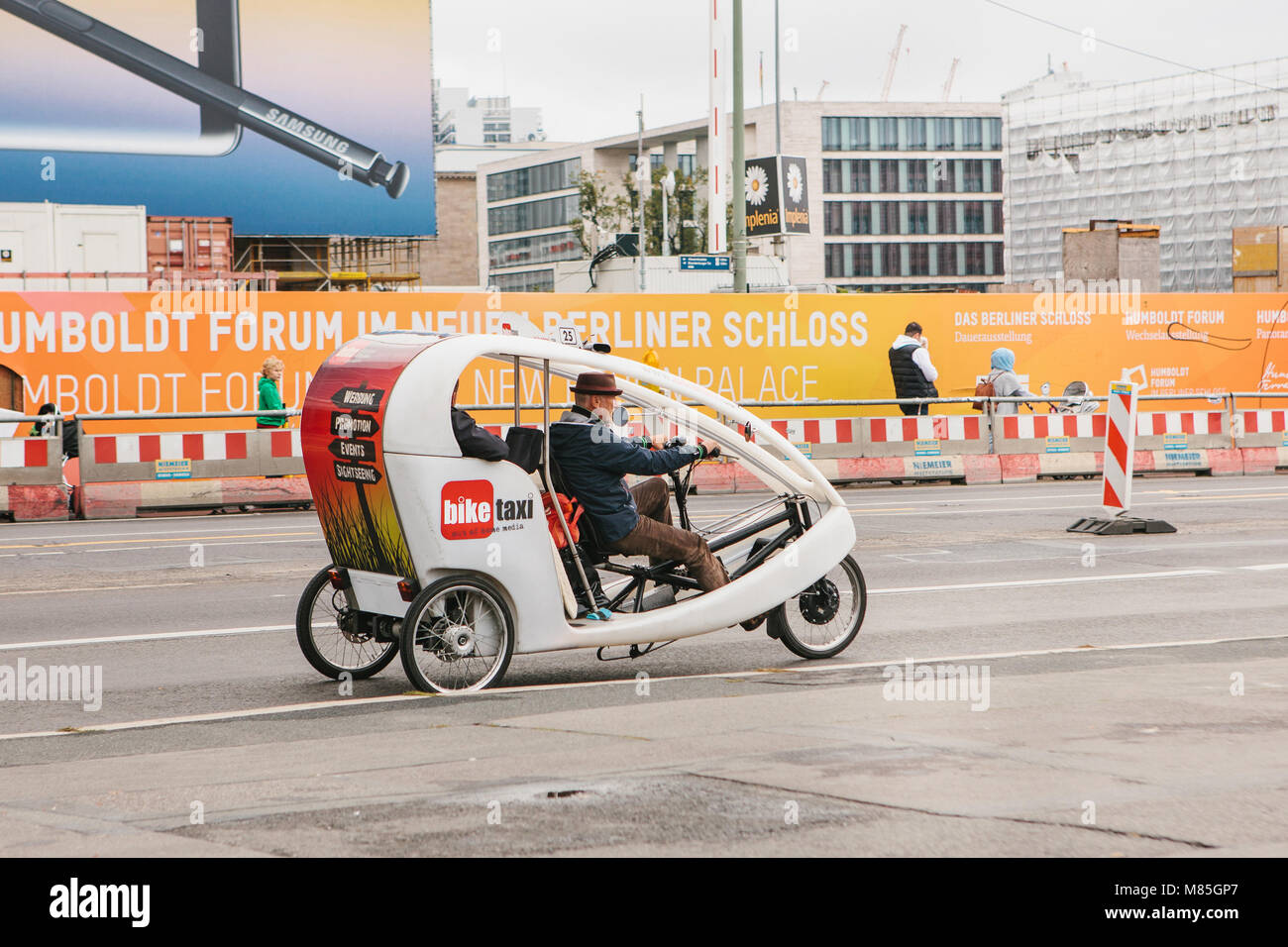 Berlin, October 1, 2017: Unknown elderly bike taxi driver carries passenger on road past people and advertising banners Stock Photo