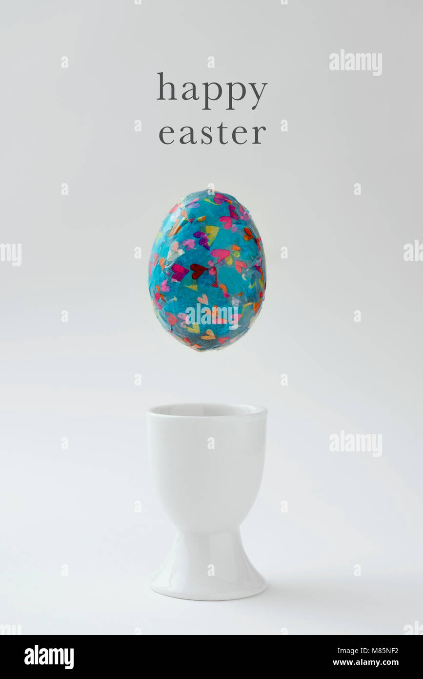 closeup of a homemade decorated easter egg, made by myself, about to fall on a white ceramic egg-cup, and the text happy easter against an off-white b Stock Photo