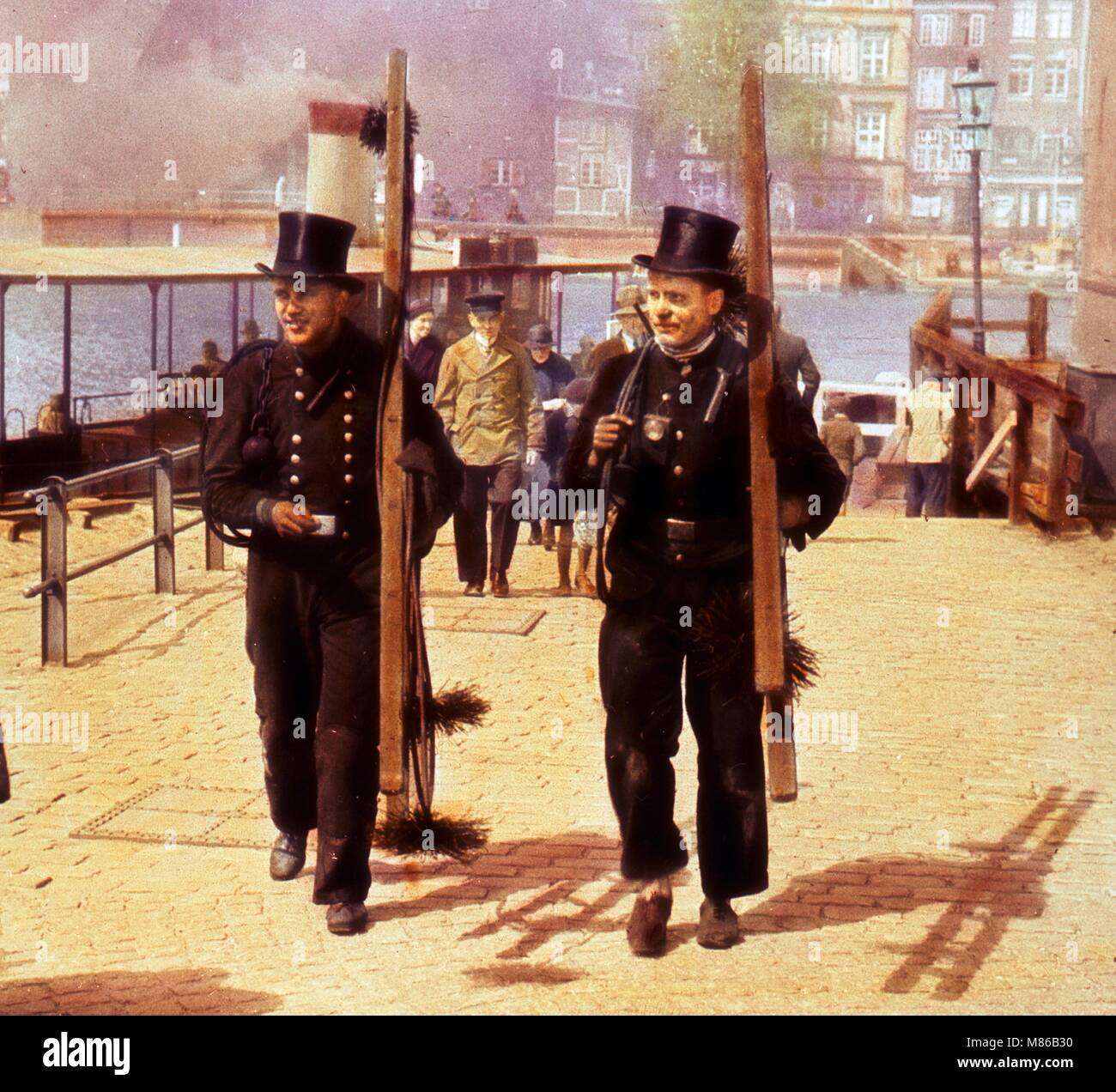 Colorized portrait of two male chimney sweeps, carrying ladders and tools, walking on a cobblestone street on the waterfront, Germany, circa 1910. (Photo by Burton Holmes) Stock Photo