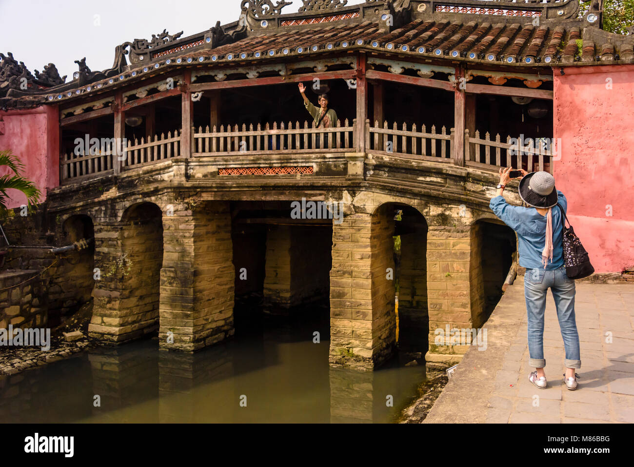 A woman takes a photograph of her husband on the Chùa Cầu Japanese bridge, an 18th century carved wooden bridge which incorporates a shrine, in Hoi An, Vietnam Stock Photo