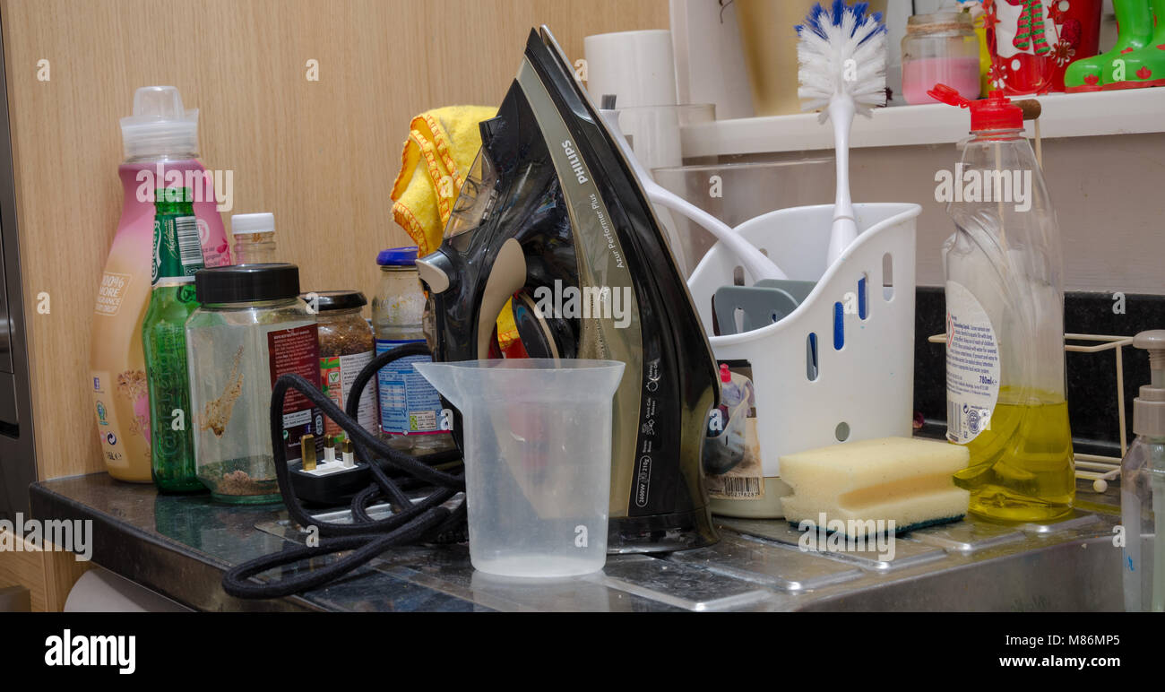 A kitchen draining board cluttered with an iron, cleaning materials and empty bottles and jars. Stock Photo