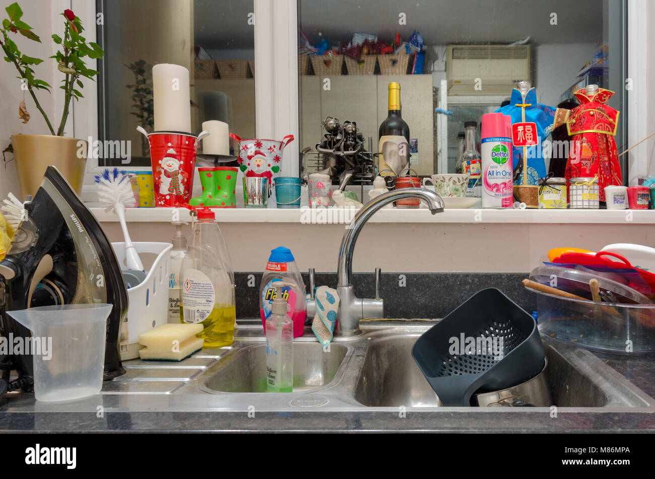 A kitchen sink cluttered with washing up, an iron, cleaning materials. Stock Photo