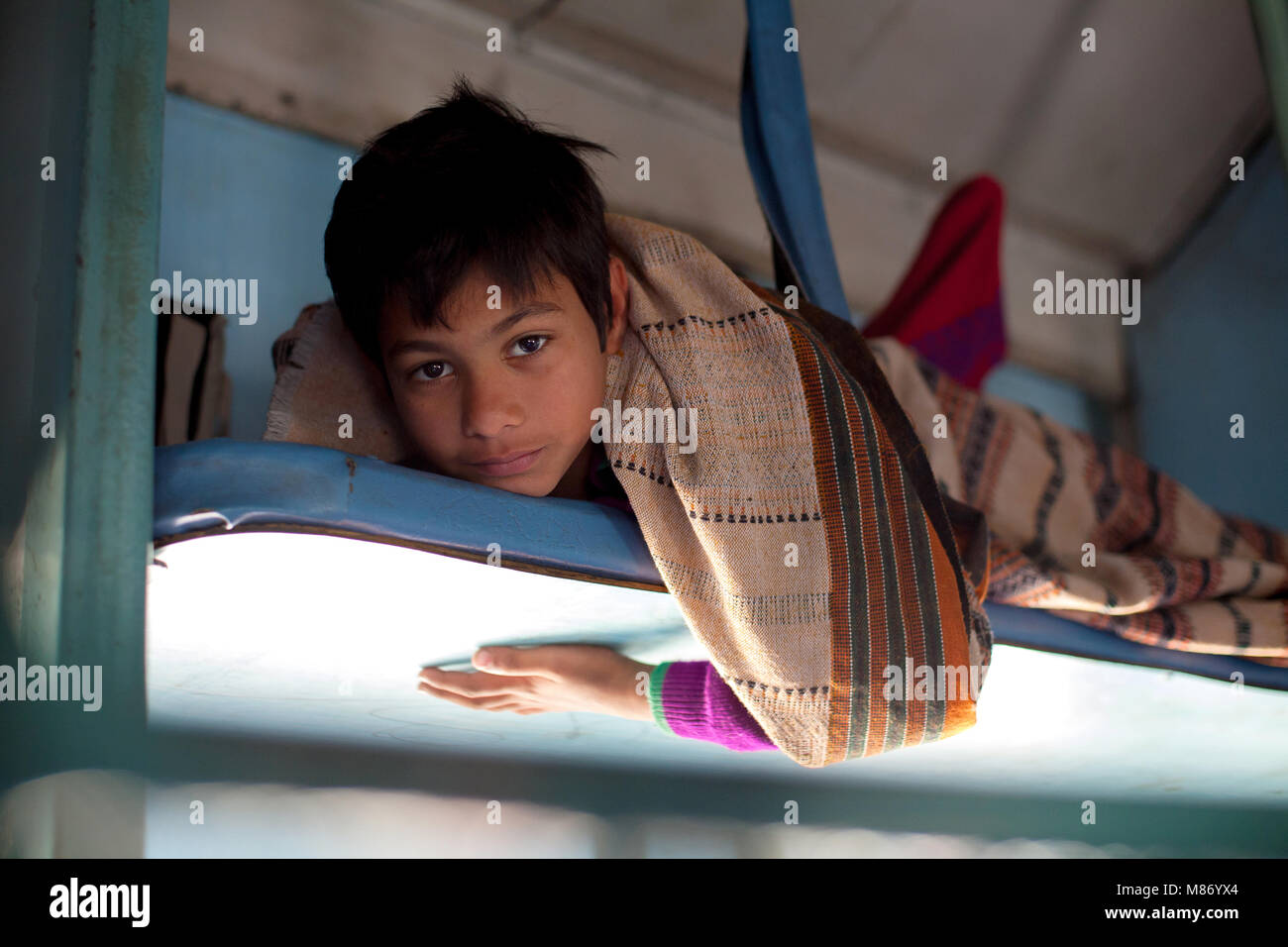 Young Indian boy laying on overhead department, traveling in train looking to camera. He is wearing purple sweather and he is covered whith nice thin  Stock Photo