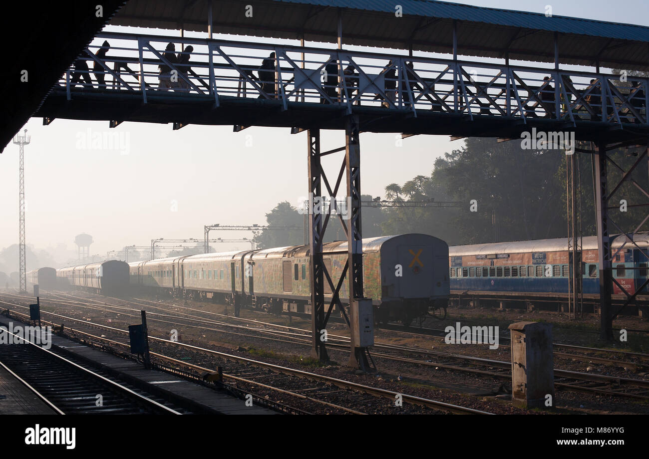 View of train station, tracks and parked train in India. People are passing over crossing bridge