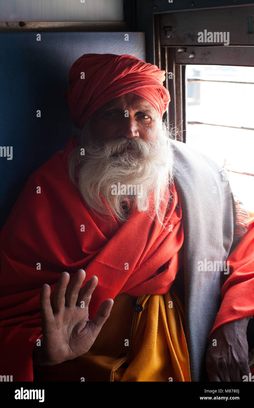Portrait of an old Indian holy man - sadhu traveling in train, sitting next to window sending his blessings with his right hand. He is wearing orange  Stock Photo
