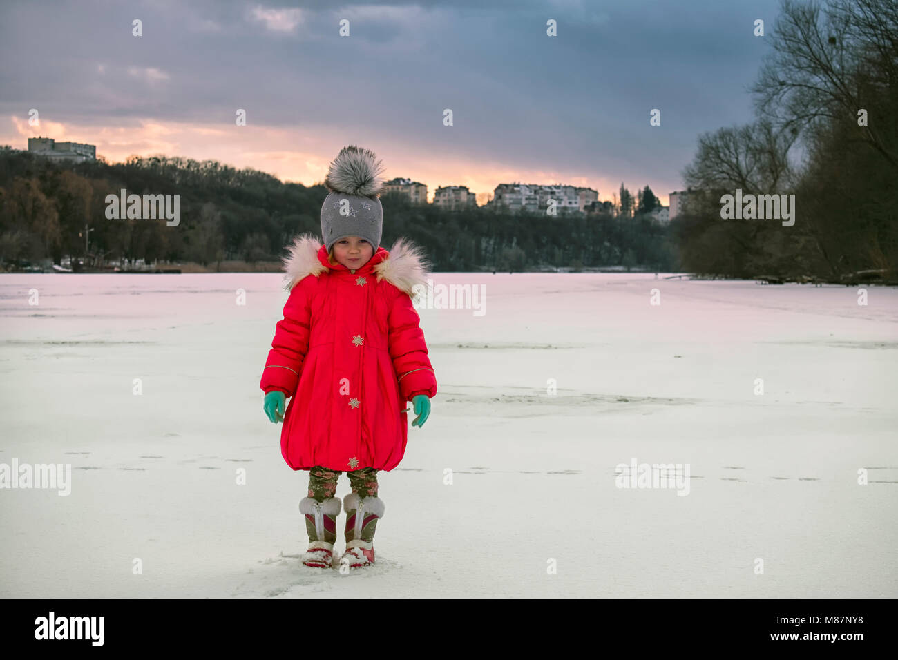 Little girl in a red down jacket stands in the middle of a frozen river