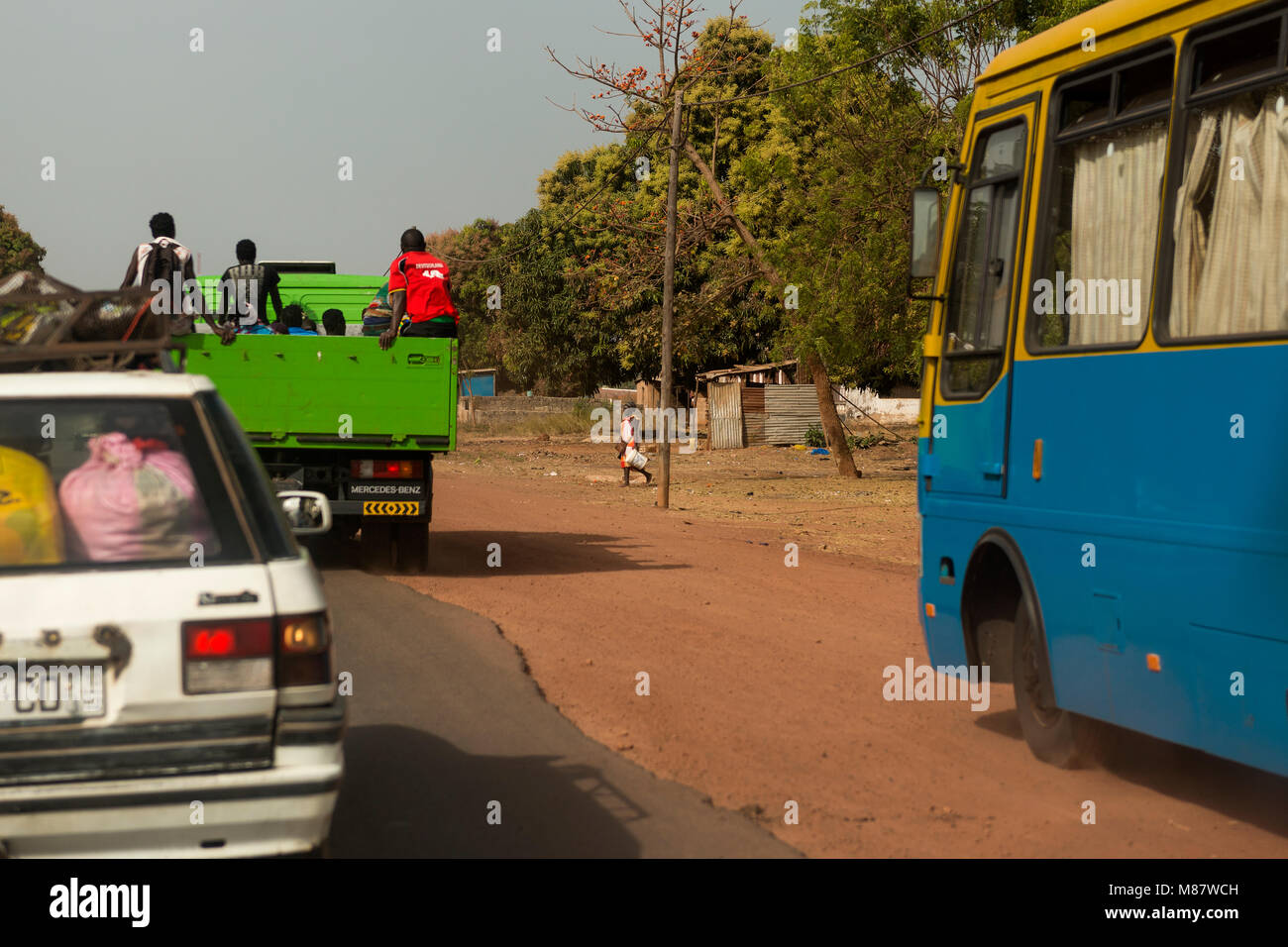 Bissau, Republic of Guinea-Bissau - January 28, 2018: Woman walking along a road with an old car and a trucky carrying people passing by her in the ou Stock Photo