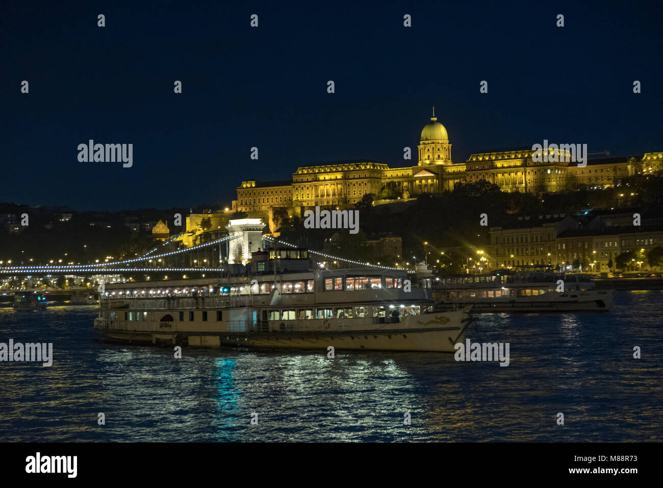 Static twilight shot of Budapest Castle and Chain Bridge lit up at night as a boat passes Stock Photo