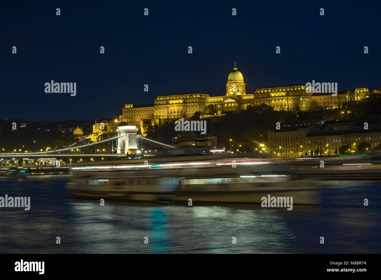 Static twilight shot of Budapest Castle and Chain Bridge lit up at night as a boat passes Stock Photo