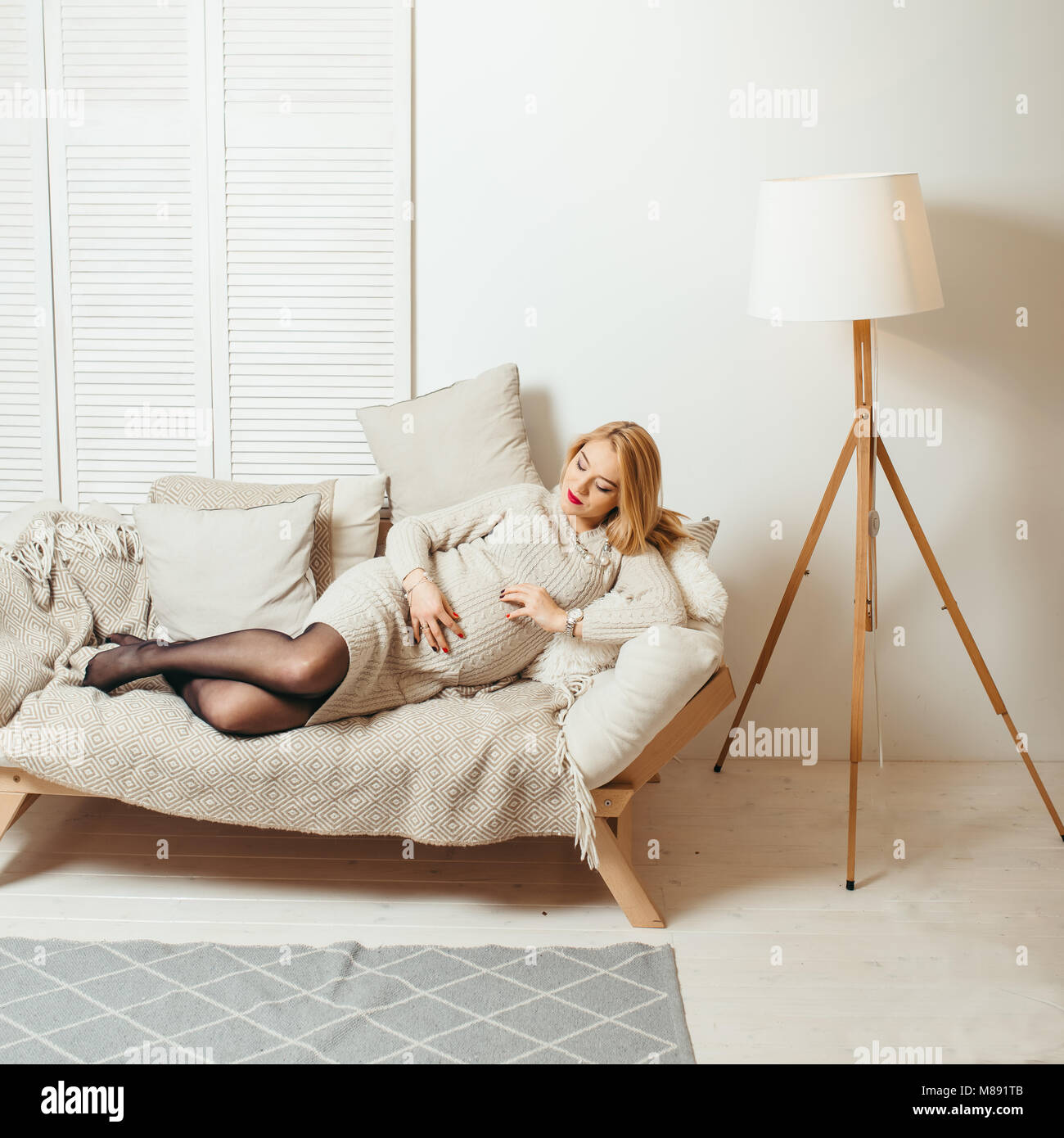 Beautiful blonde pregnant woman with red lipstick is touching her stomach and laying on the cozy sofa in the white room. Stock Photo