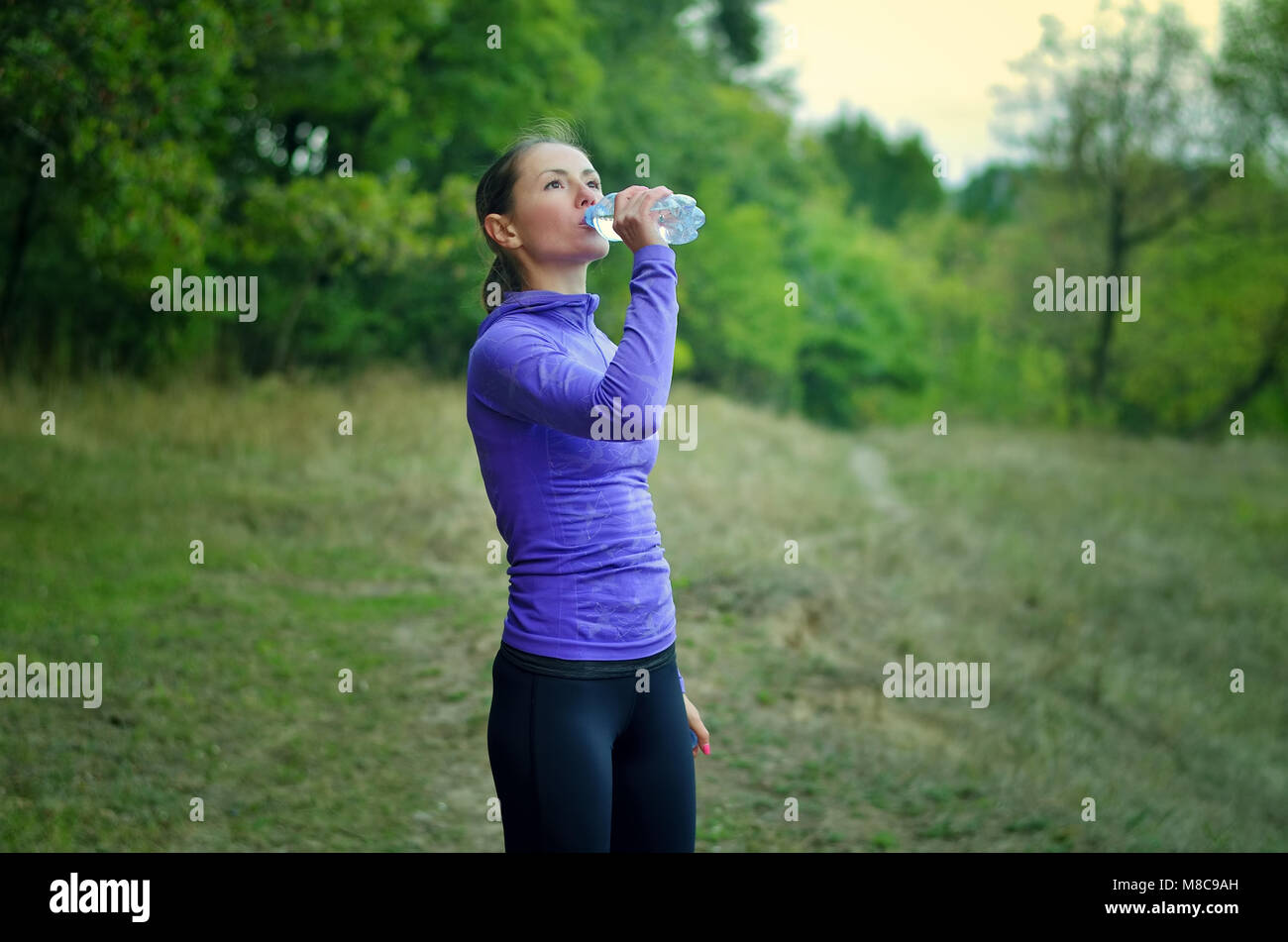 A caucasian slim woman in a blue sports jacket with a hood and black  leggins drink water from the bottle after jogging on a colorful green forest hil Stock Photo