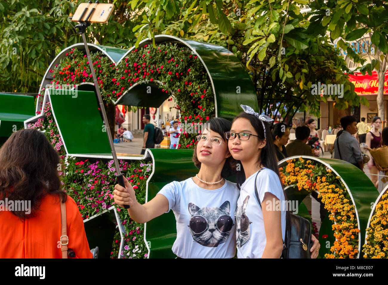 Two girls take a selfie using a selfie stick in front of Chinese Lunar New Year decorations, Ho Chi Minh City, Saigon, Vietnam Stock Photo