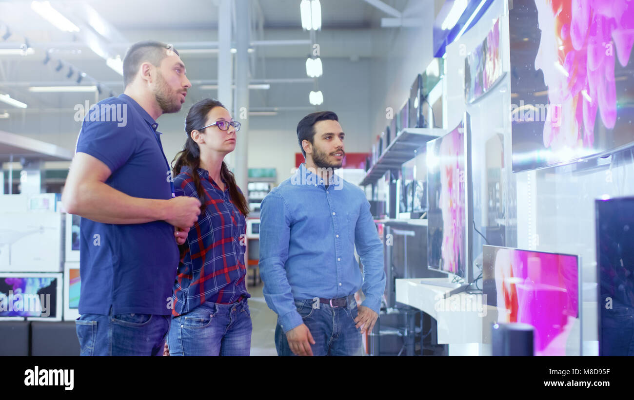In the Electronics Store Professional Consultant Approaches Young Couple with His Expert Advice on Which of New Models of 4K TV's is Best for Them Stock Photo