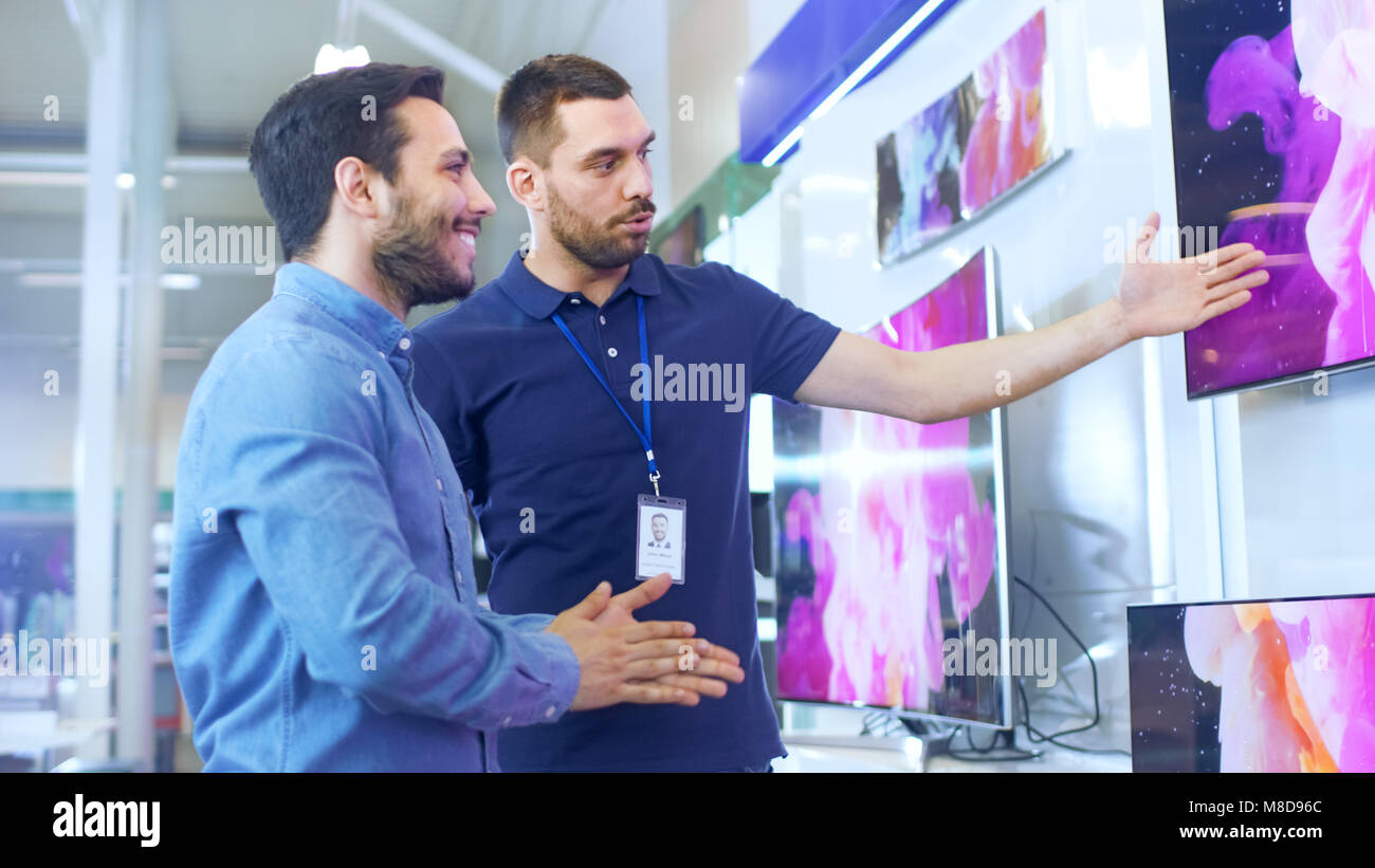 In the Electronics Store Professional Consultant Shows Latest 4K UHD TV's to a Young Man, They Talk about Specifications and What Model is Best Stock Photo