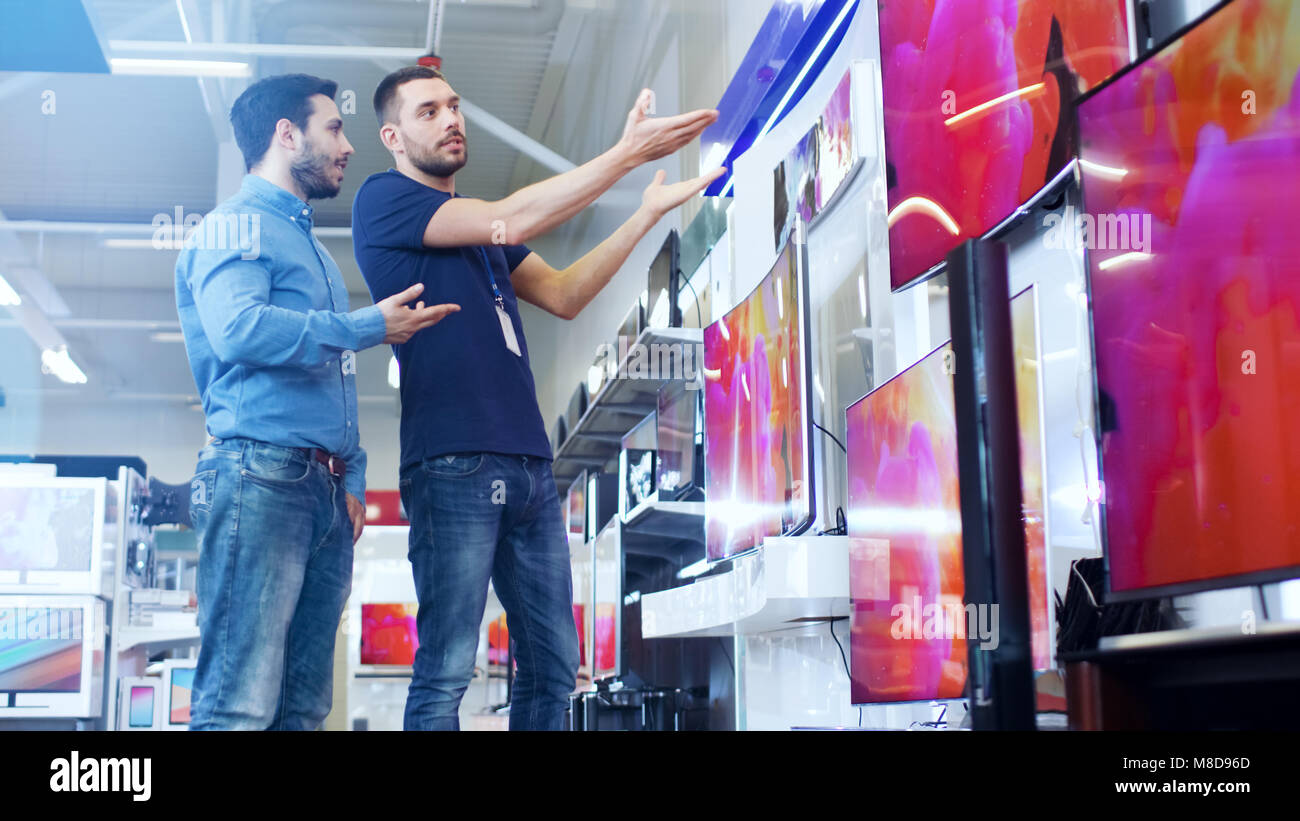 In the Electronics Store Professional Consultant Shows Latest 4K UHD TV's to a Young Man, They Talk about Specifications and What Model is Best Stock Photo