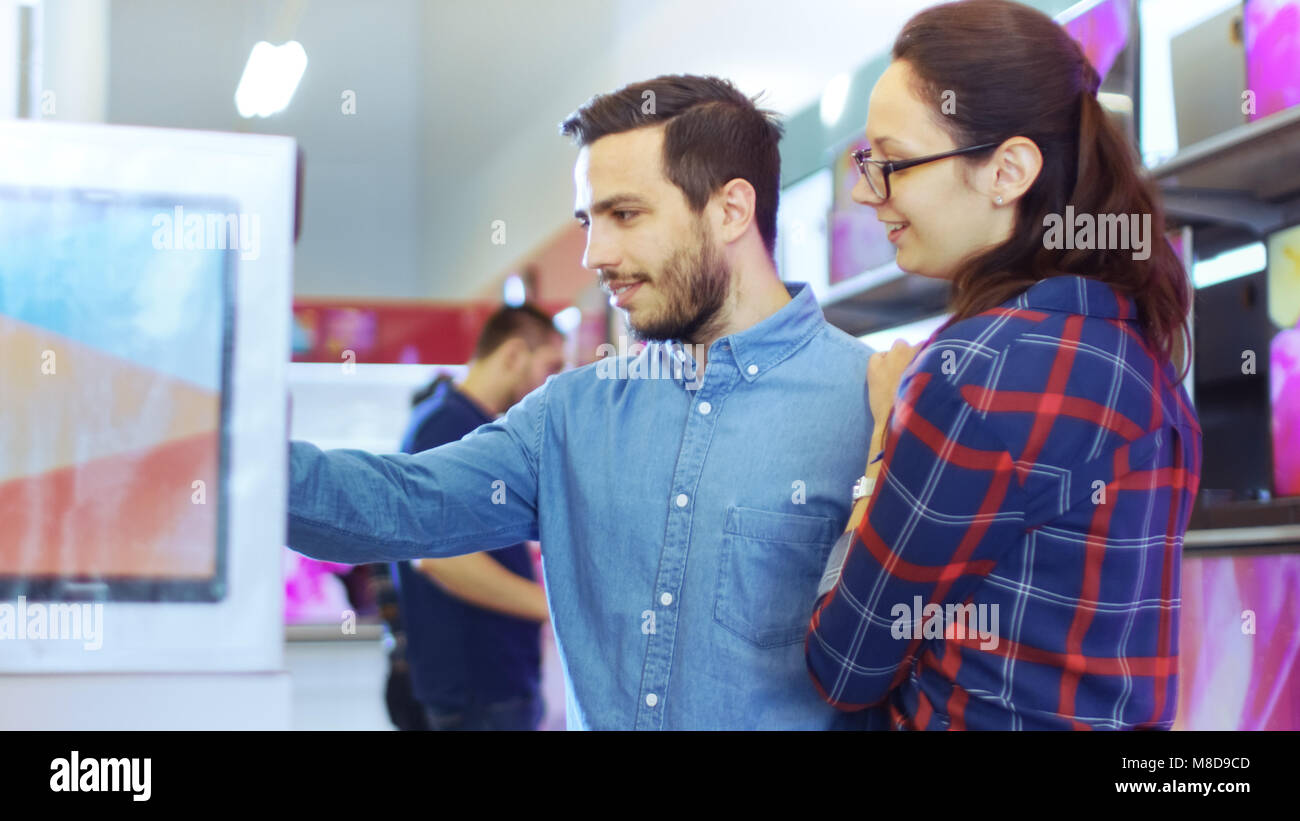 Beautiful Young Couple in the Electronics Store Browsing, Looking for Newest Gadgets, Tablets and Photo/ Video Cameras Presented on the Shelves. Stock Photo