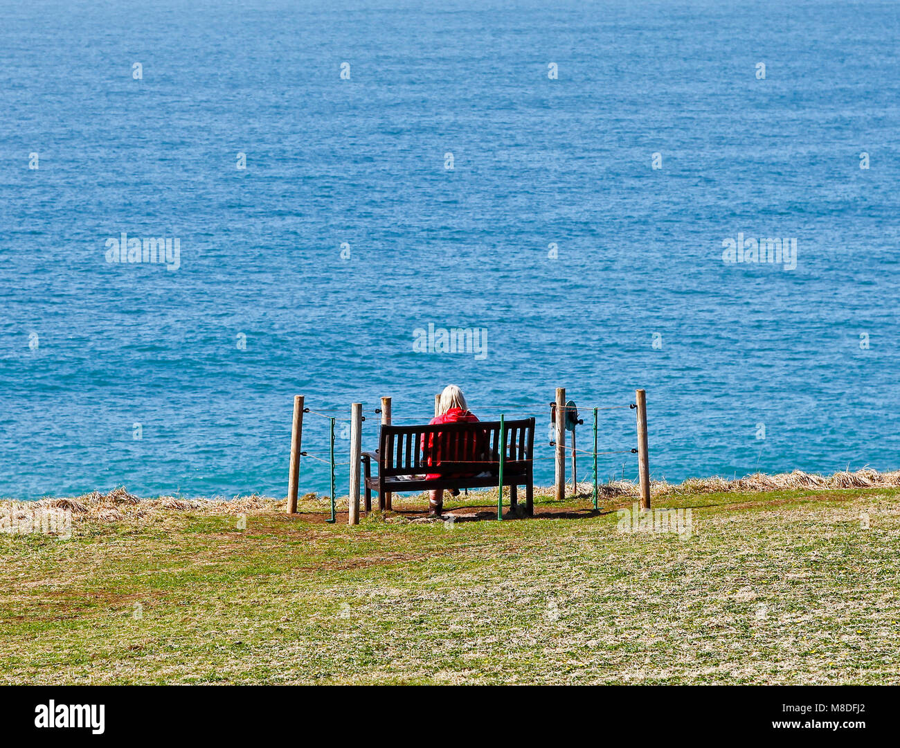 A woman sitting on an isolated bench looking out to sea on the Lizard ...