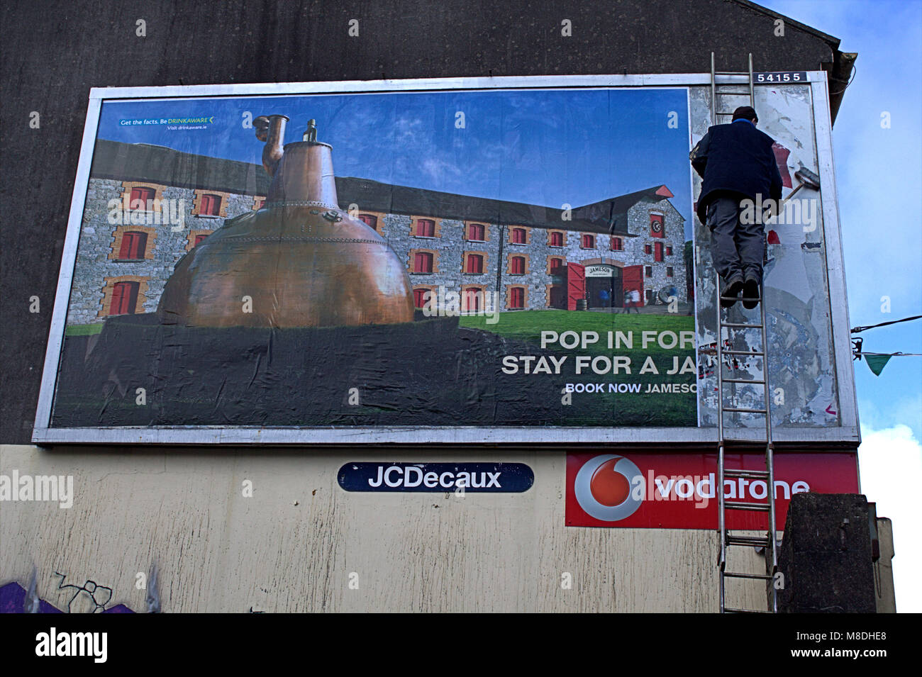 man up a ladder putting up a new advertising hoarding, billboard of jamesons whisky a famous irish whisky brand. skibbereen, ireland Stock Photo