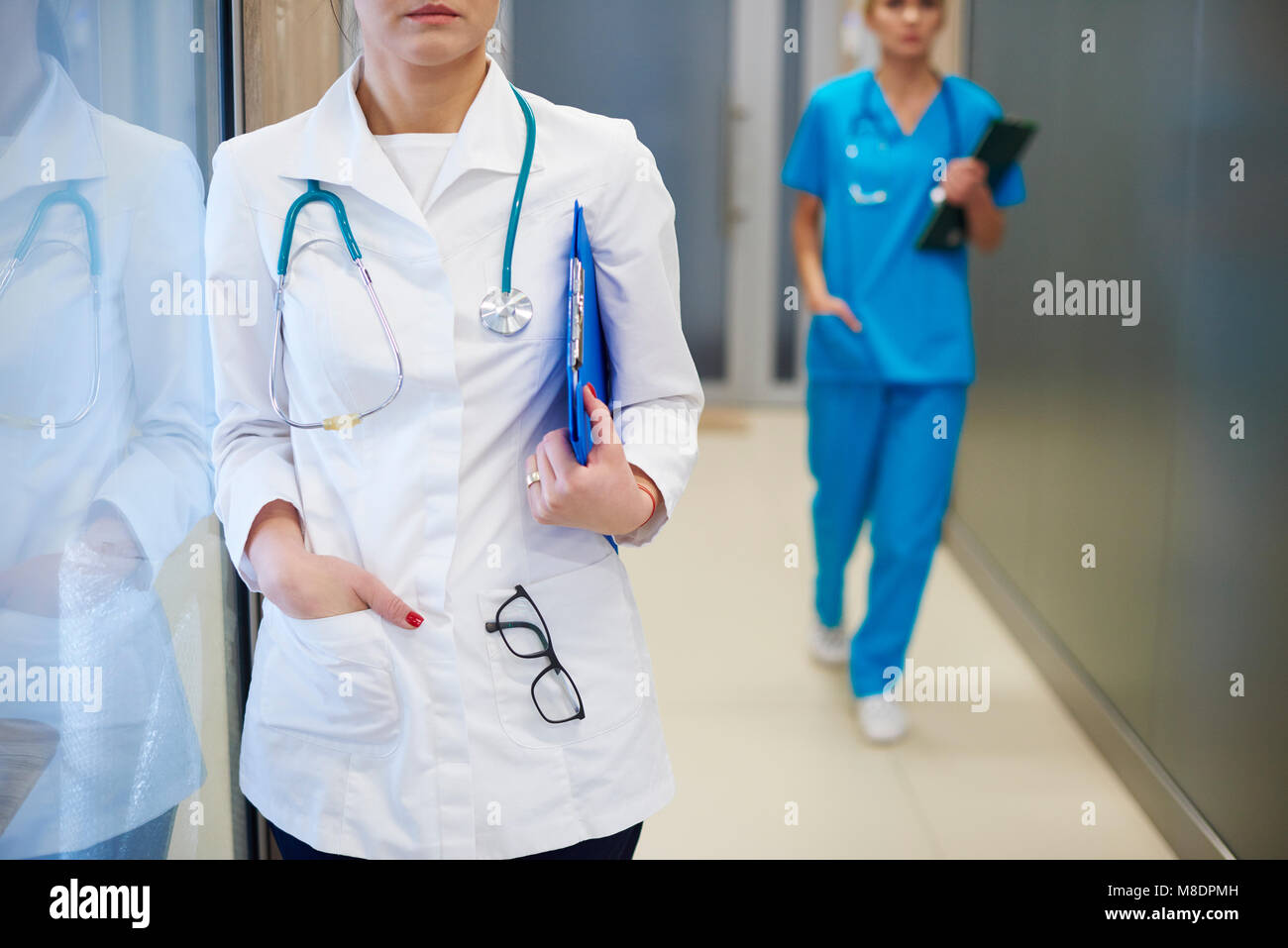 Doctor standing in hospital hallway, surgeon walking in background, mid section Stock Photo