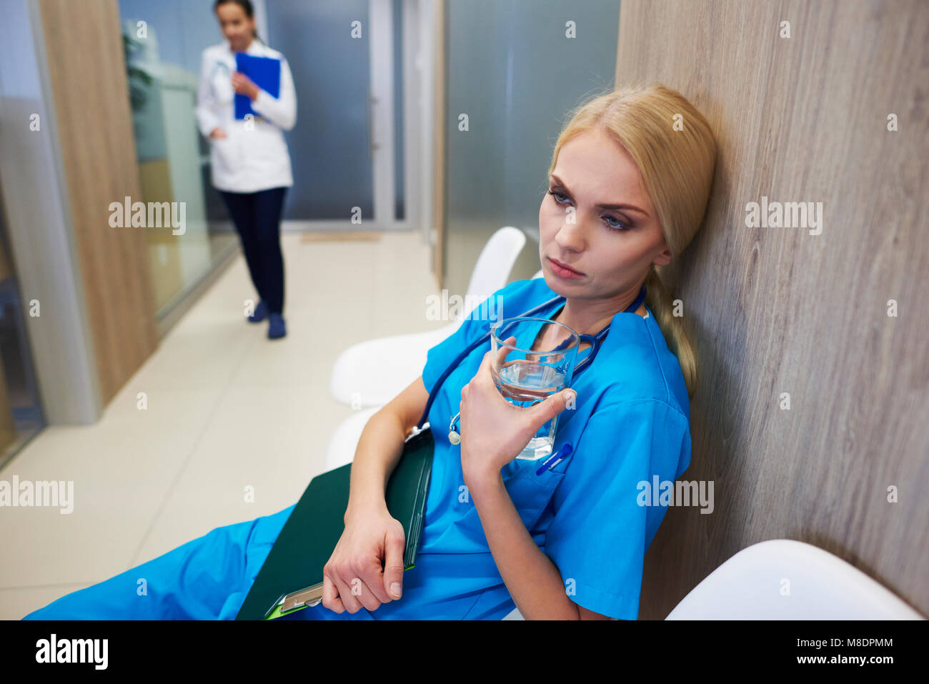 Surgeon sitting in hospital hallway, holding glass of water, pensive expression Stock Photo